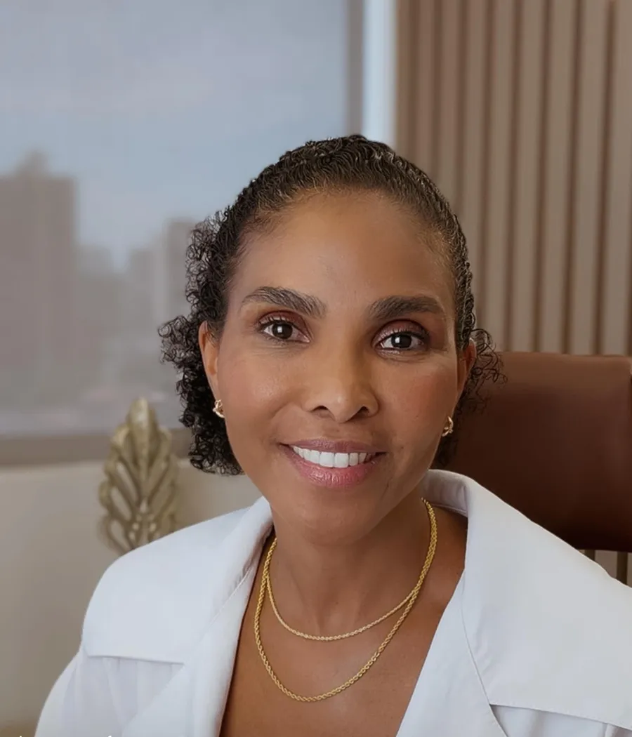 Dra. Rosana Frempong sentada à mesa do consultório, de jaleco branco, sorrindo